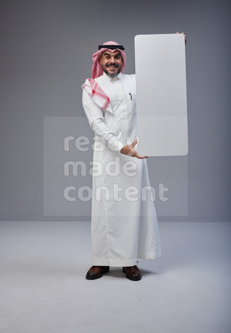 Saudi man Wearing Thob and red Shomag standing holding board on Gray background