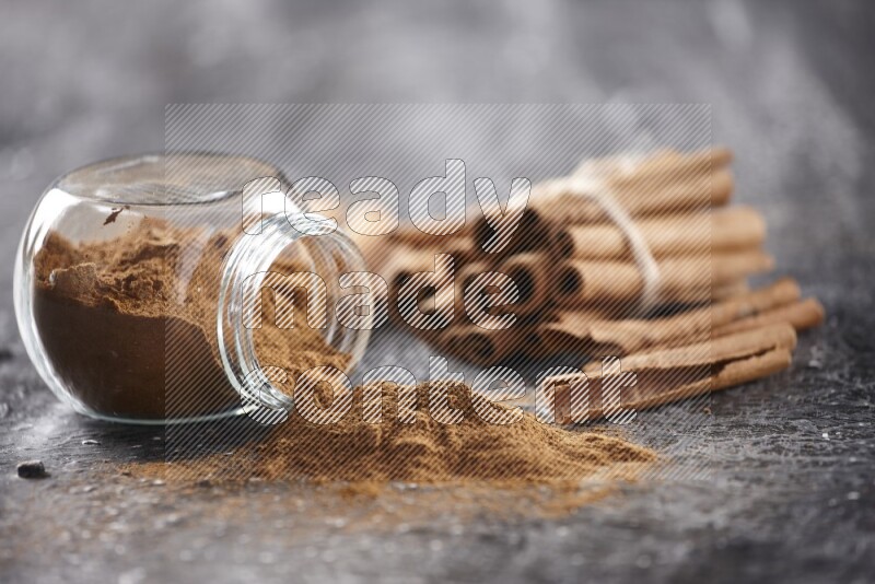 Herbal glass jar full of cinnamon powder flipped with cinnamon sticks stacked and bounded on a textured black background