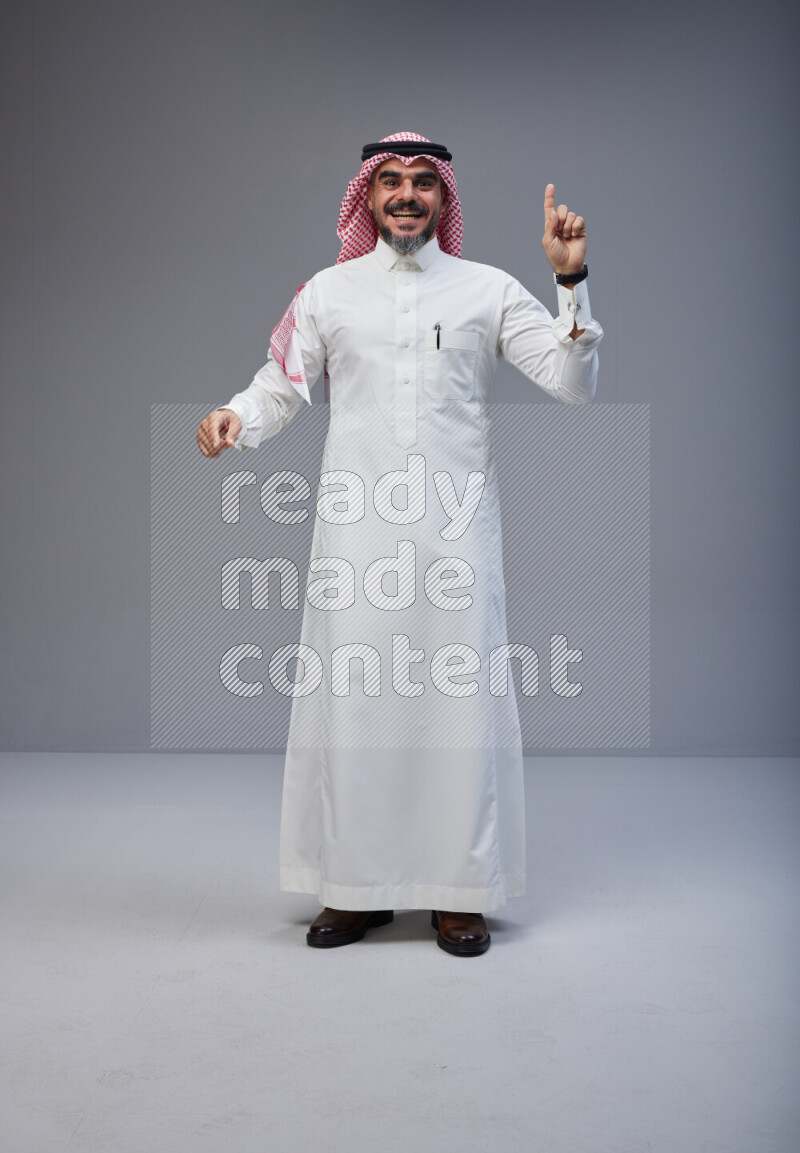 Saudi man Wearing Thob and red white Shomag standing interacting with the camera on Gray background