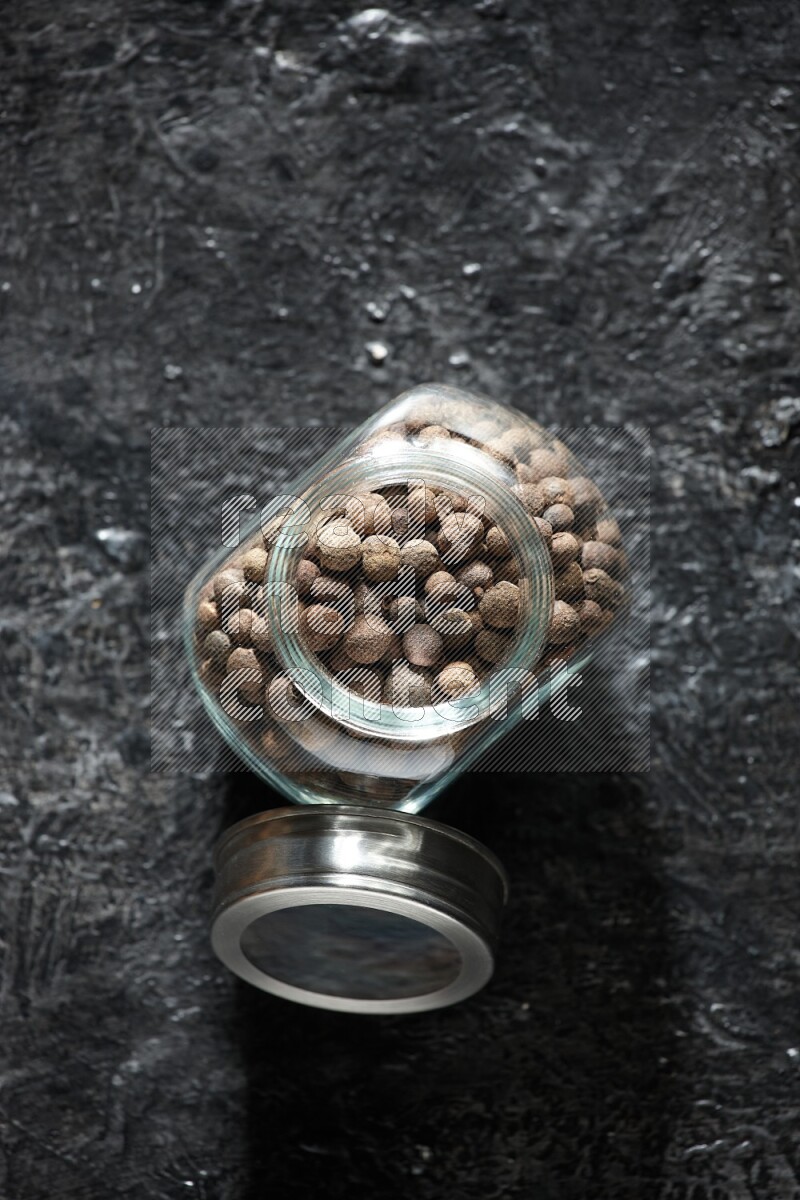 A glass spice jar full of allspice whole balls on a textured black flooring
