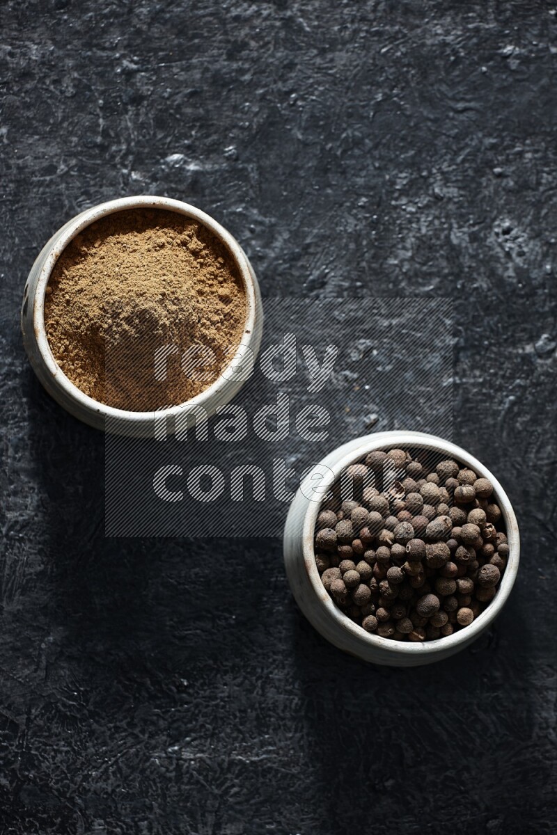 2 beige bowls, one full of allspice powder and the other full of whole balls on a textured black flooring