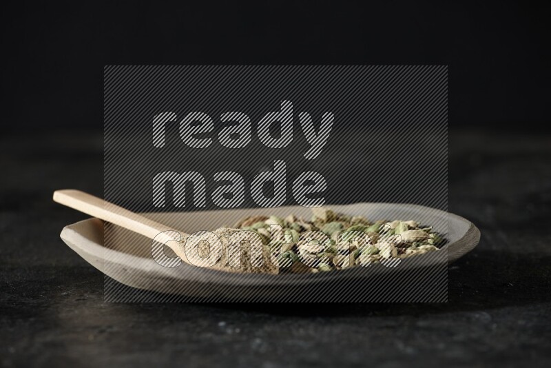 A plate filled with cardamom seeds and a wooden spoon full of cardamom powder on a textured black flooring