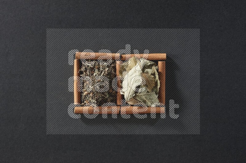 مربعان من أعواد القرفة ممتلئان بأوراق الغار والريحان المجفف علي خلفية سوداء