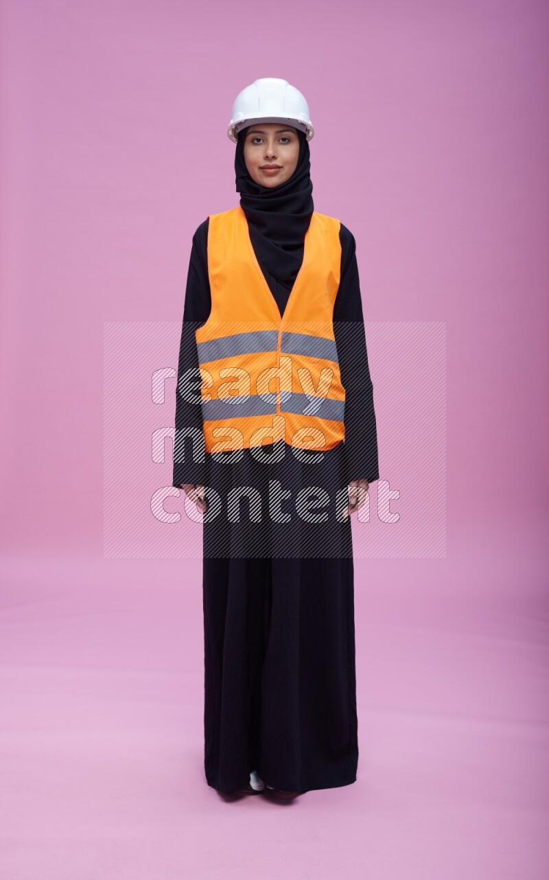 Saudi woman wearing Abaya with engineer vest and helmet standing interacting with the camera on pink background