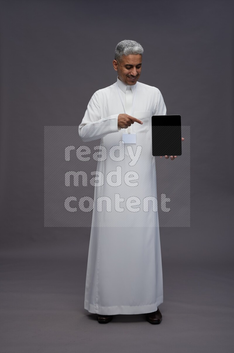 Saudi man wearing thob with neck strap employee badge standing showing tablet to camera on gray background