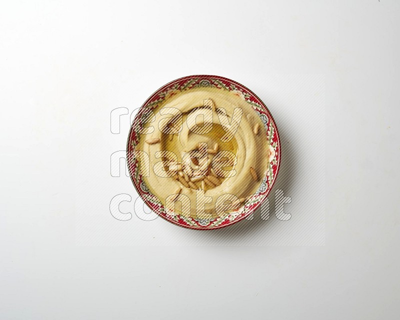 Hummus in a red plate with patterns garnished with pine nuts on a white background