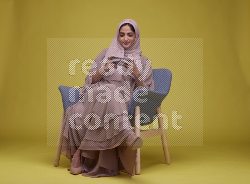 A woman Playing Game Sitting  on a Yellow Background wearing Brown Abaya with Hijab