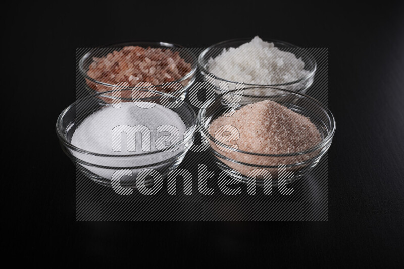 4 glass bowls filled with table salt, coarse sea salt, fine himalayan salt and coarse himalayan salt on black background