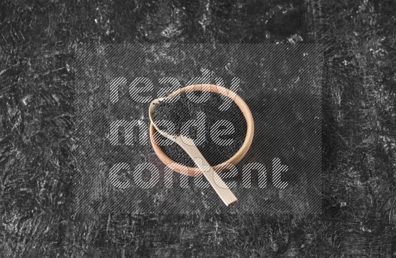 A wooden bowl full of black seeds with wooden spoon full of the seeds on it on a textured black flooring
