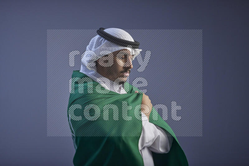 A close-up shot of Saudi man wearing thob and white shomag wrapping big Saudi flag on gray background