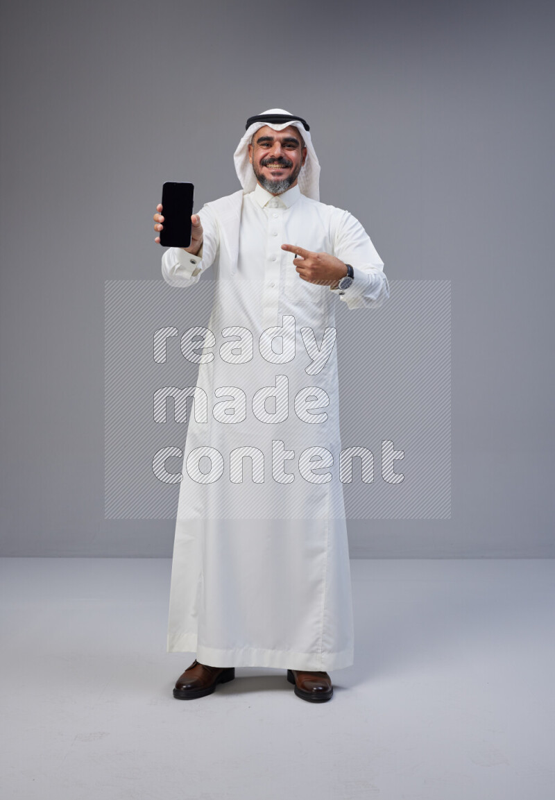 Saudi man Wearing Thob and white Shomag standing showing phone to camera on Gray background
