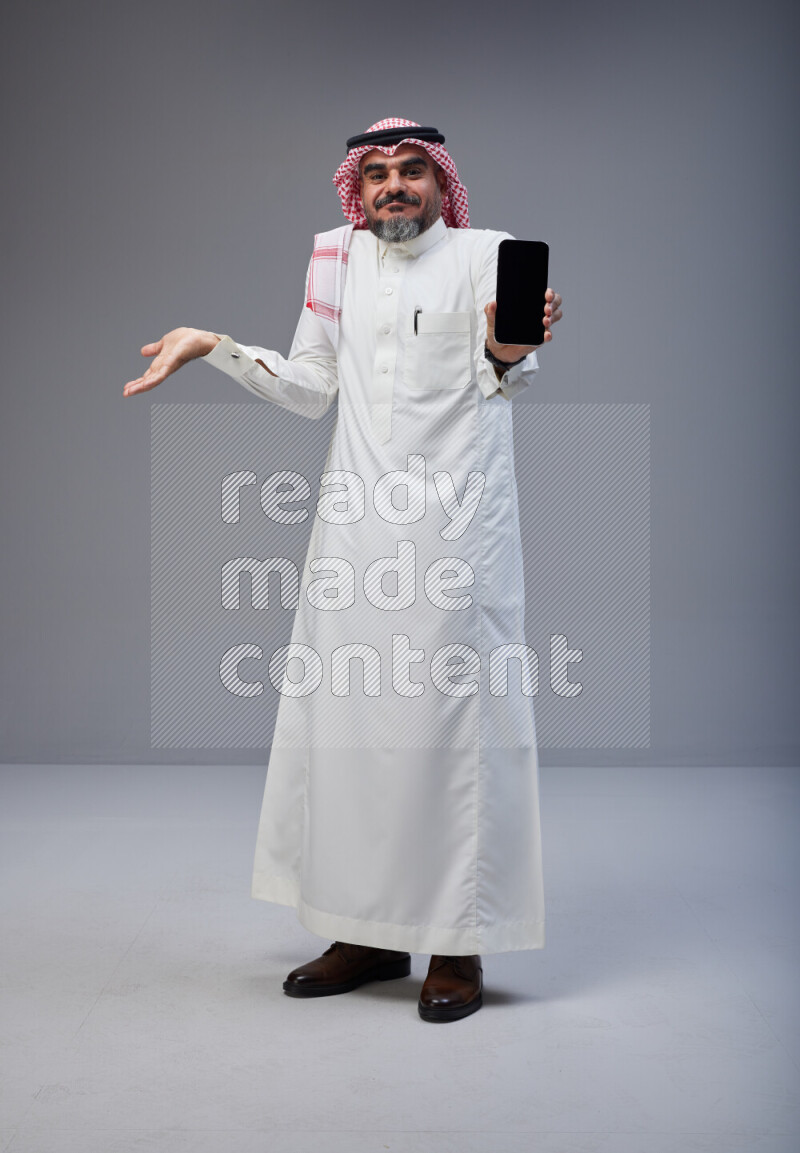 Saudi man Wearing Thob and red Shomag standing showing phone to camera on Gray background