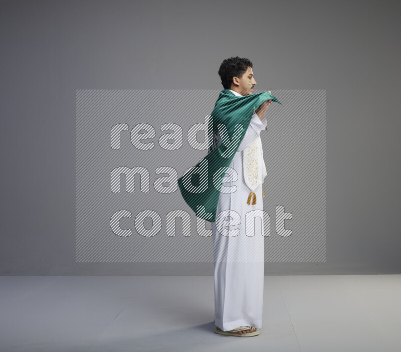 A Saudi man standing wearing thob with face painting wrapping Saudi flag scarf and holding big Saudi flag on gray background