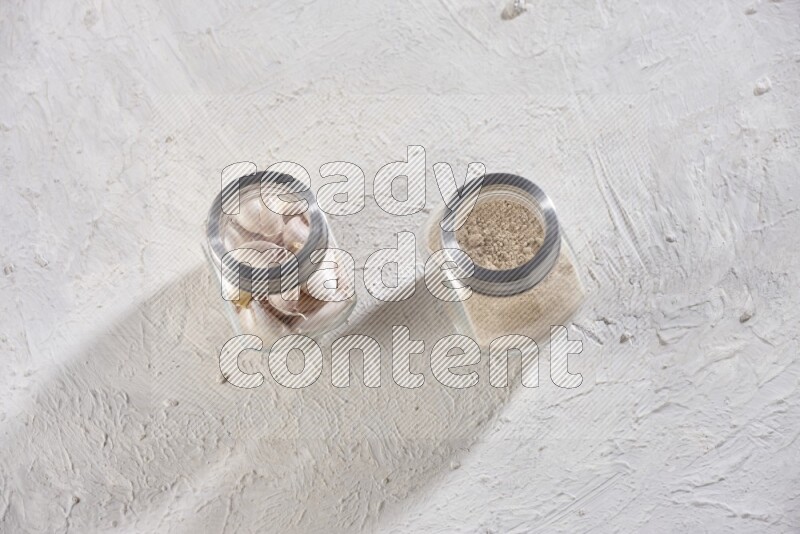 2 glass spice jars one full of garlic powder and the other full of cloves on a textured white flooring