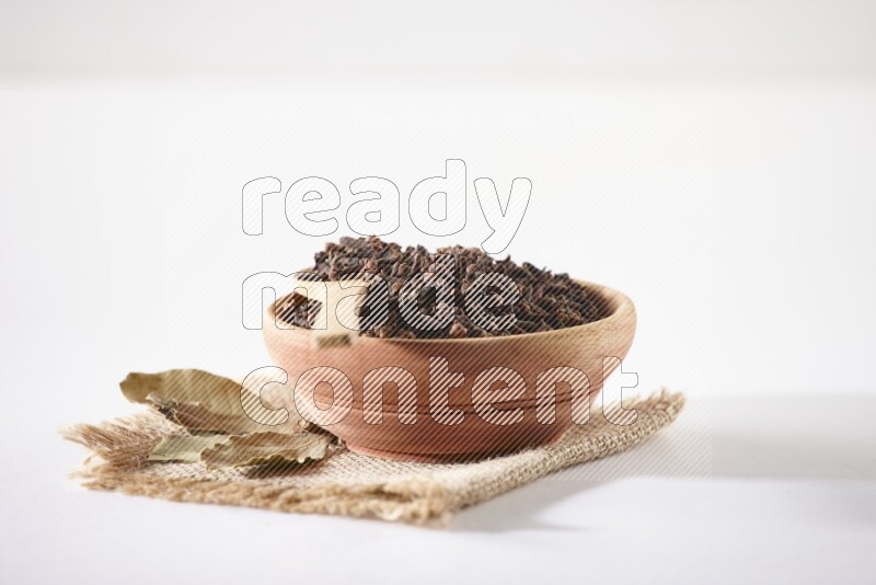 A wooden bowl and a wooden spoon full of cloves on a piece of burlap on a white flooring