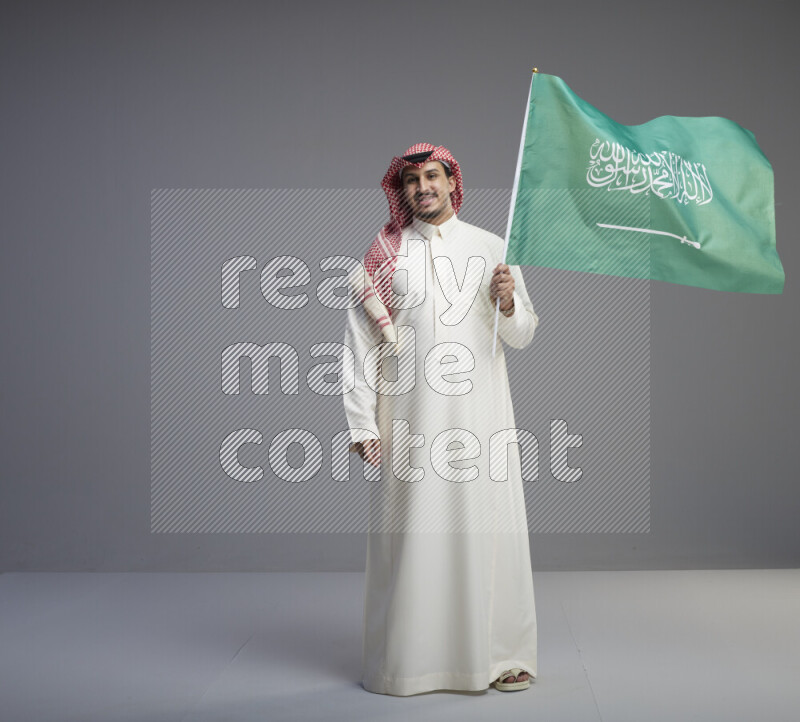 A Saudi man standing wearing thob and red shomag raising big Saudi flag on gray background