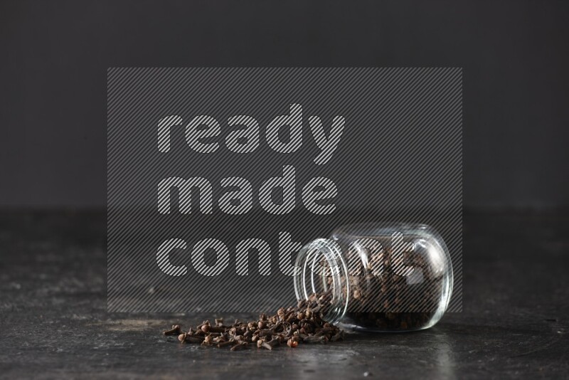 وعاء زجاجي للبهارات مقلوب ممتلئ بالقرنفل والقرنفل سقط منه على أرضية سوداء