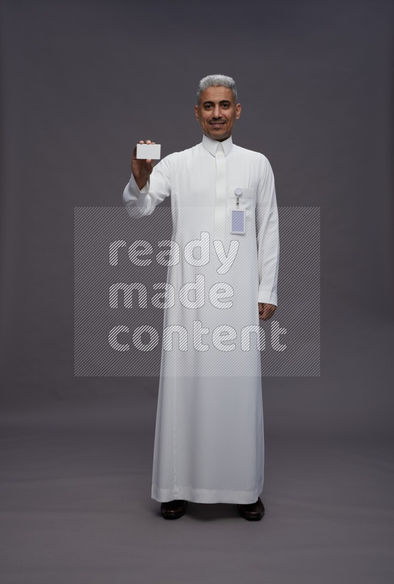 Saudi man wearing thob with pocket employee badge standing holding ATM card on gray background