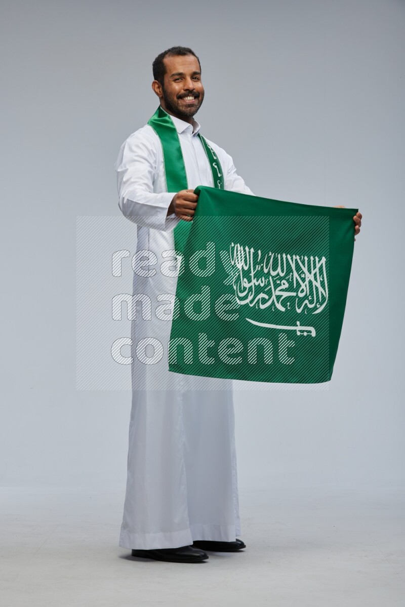 Saudi man wearing Thob standing holding Saudi flag on Gray background