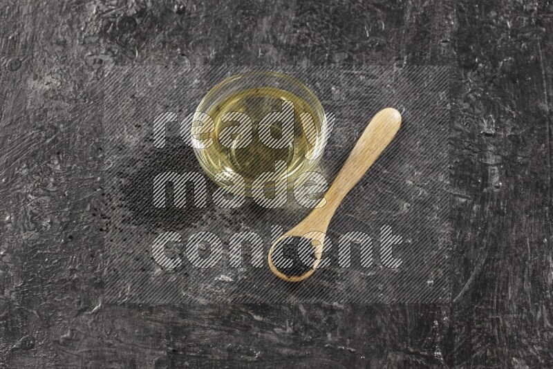 A glass bowl full of black seeds oil and wooden spoon full of black seeds with seeds spreaded on a textured black flooring