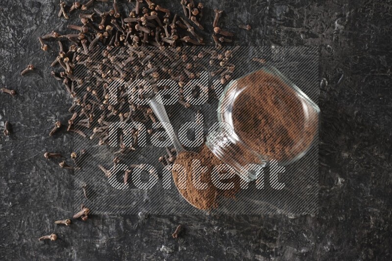 A flipped glass spice jar and a metal spoon full of cloves powder with cloves spread on textured black flooring