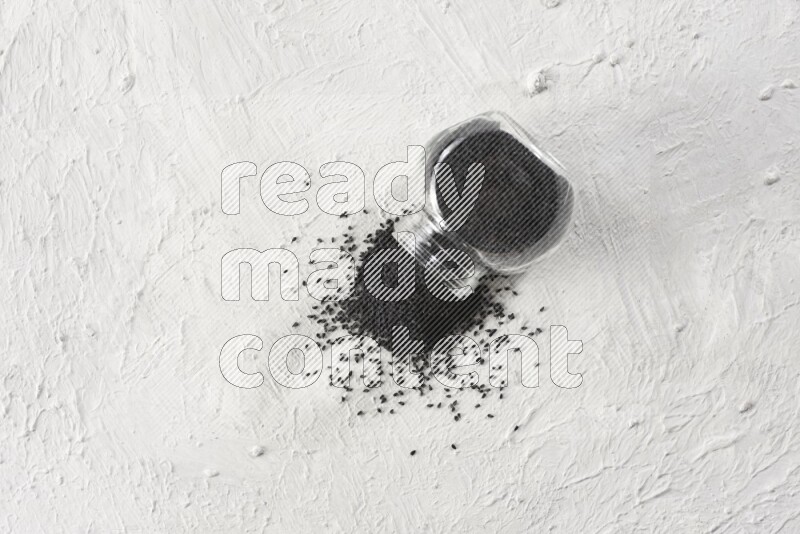 A glass spice jar full of black seeds and the jar flipped and seeds spread on a textured white flooring
