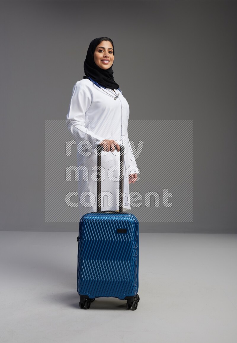 Saudi woman wearing lab coat with stethoscope standing holding Travel bag on Gray background