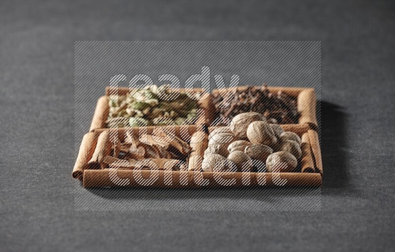 4 مربعات من عيدان القرفة ممتلئة بقطع القرفة وحبوب الهيل والقرنفل وجوزة الطيب على أرضية سوداء