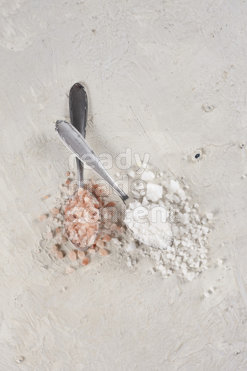 2 metal spoons filled with white salt and pink himalayan salt on white background
