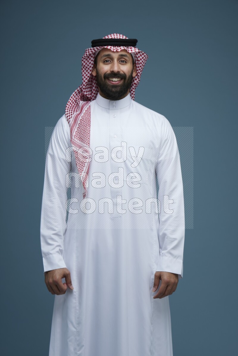 A man posing in a blue background wearing Saudi Thob and Shomag