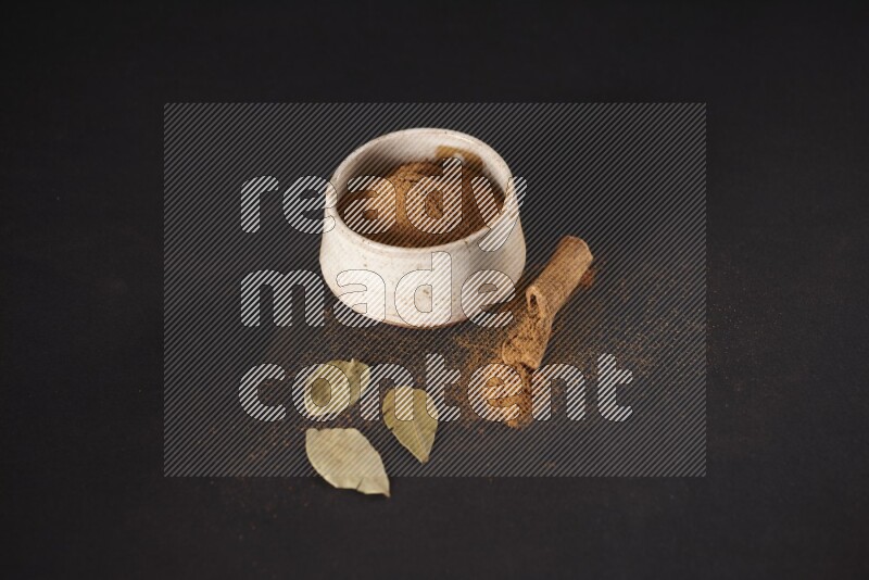 Cinnamon powder in a white pottery bowl and cinnamon sticks and laurel leaves on black background
