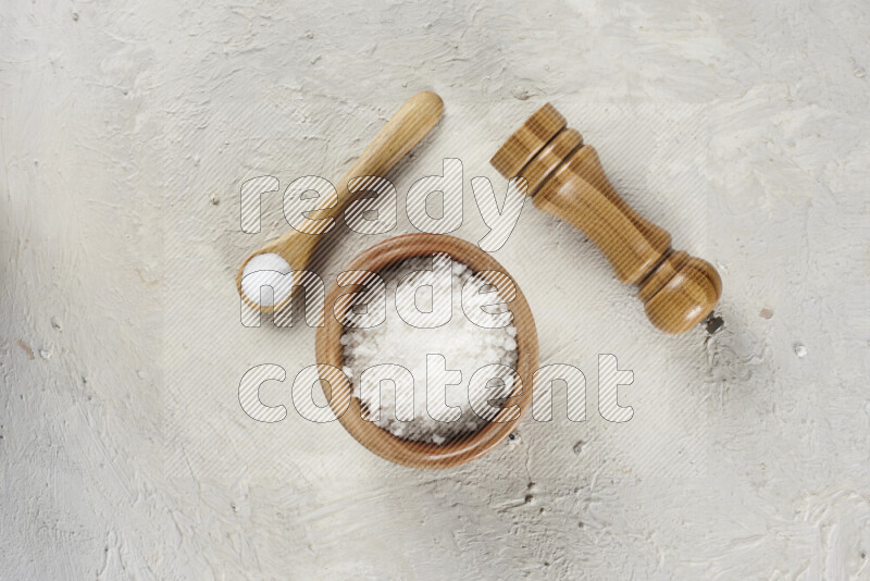 A wooden bowl and spoon filled with white sea salt and wooden grinder beside them on white background