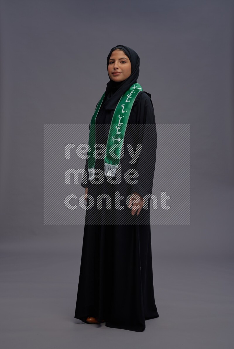 Saudi woman wearing Abaya standing holding Saudi flag on gray background