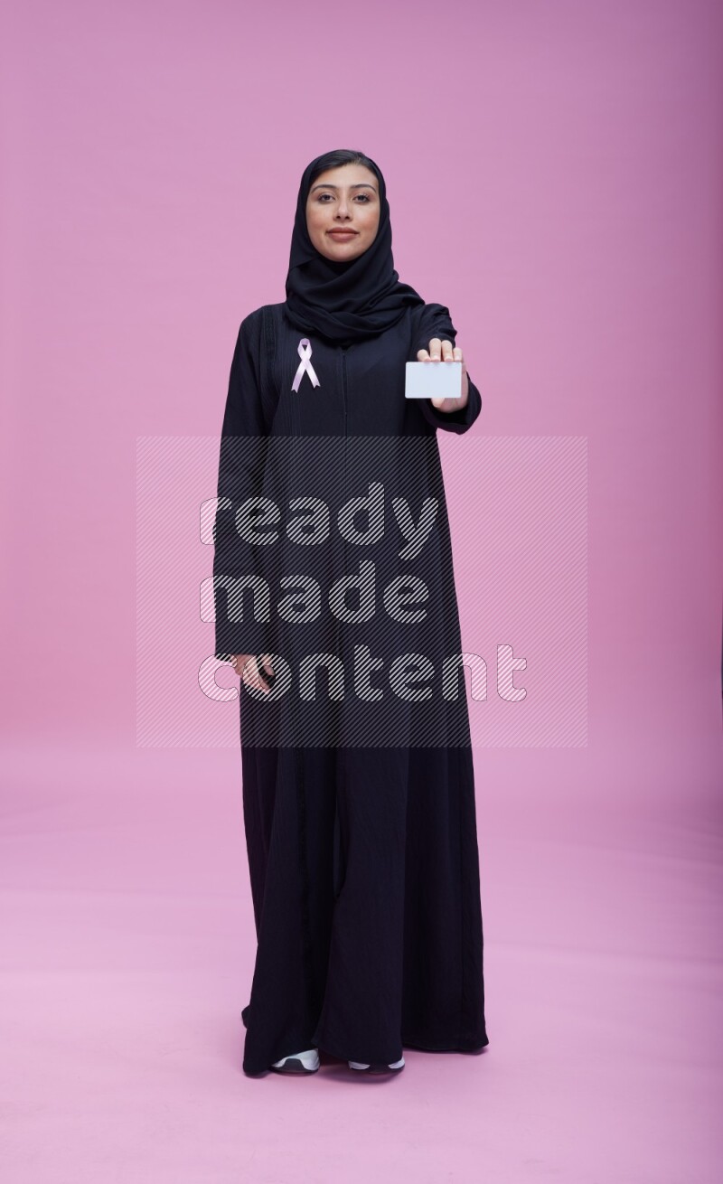 Saudi woman wearing pink ribbon on Abaya standing holding ATM card on pink background