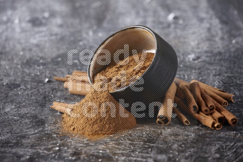 Black pottery bowl over filled with cinnamon powder and cinnamon sticks around the bowl on a textured black background