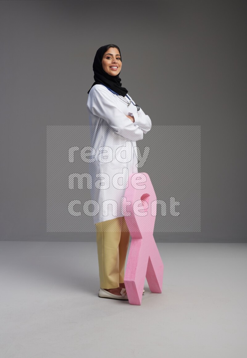 Saudi woman wearing lab coat with stethoscope standing holding awareness ribbon symbol standing on Gray background