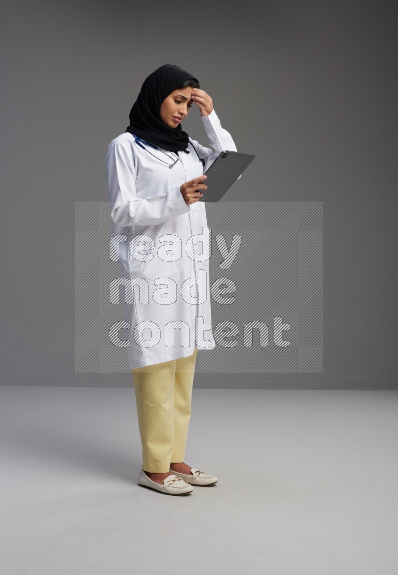 Saudi woman wearing lab coat with stethoscope standing working on tablet on Gray background