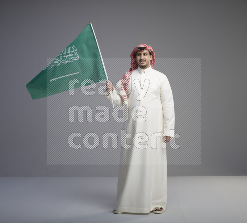 A Saudi man standing wearing thob and red shomag raising big Saudi flag on gray background