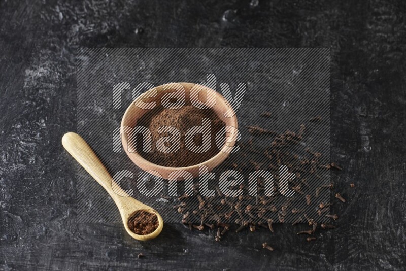 A wooden bowl and wooden spoon full of cloves powder with spreaded cloves on textured black flooring