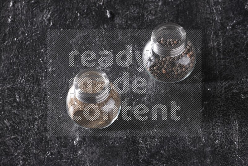 2 glass spice jars full of black pepper powder and black pepper beads on textured black flooring