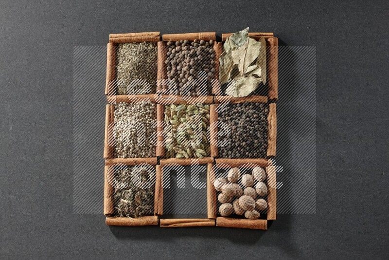 9 مربعات من أعواد القرفة ممتلئة بالهيل في المنتصف محاطة بجوزة الطيب والفلفل الأسود وورق الغار والبهارات والكمون والفلفل الأبيض والريحان المجفف والملح على أرضية سوداء