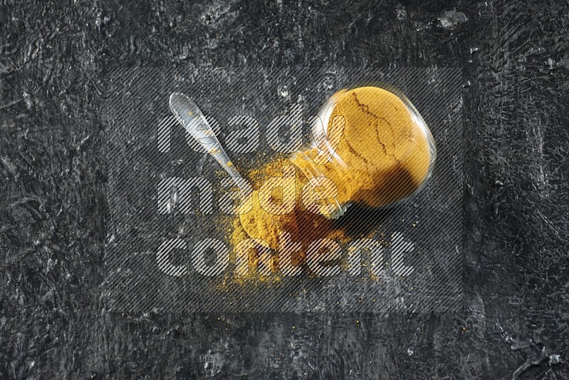 وعاء زجاجي للتوابل مقلوب وملعقة معدنية ممتلئان ببودرة الكركم والبودرة تناثرت منه على خلفية سوداء