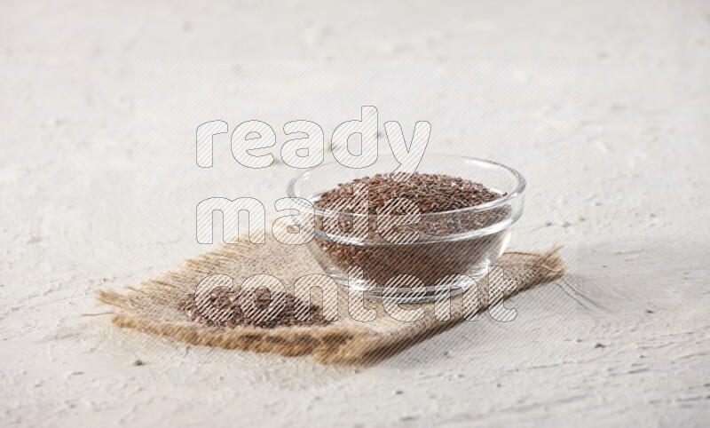 A glass bowl full of flax seeds and a bunch of seeds on burlap fabric on a textured white flooring