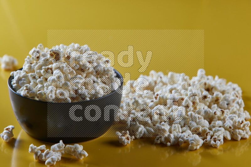 A black ceramic bowl full of popcorn with popcorn beside it on a yellow background in different angles