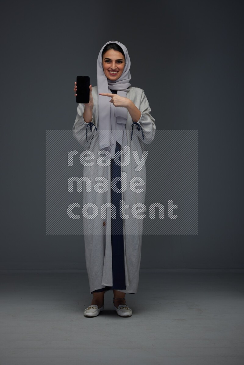A Saudi woman wearing a light gray Abaya and head scarf standing and showing the phone's screen while holding a blue luggage eye level on a grey background