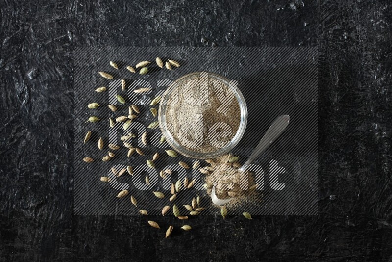 A glass bowl and a metal spoon full of cardamom powder with cardamom seeds beside them on textured black flooring