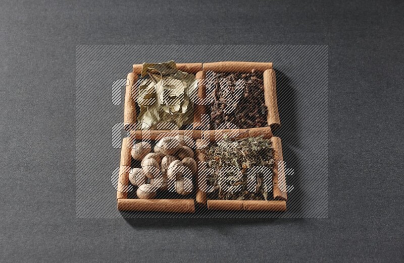 4 مربعات من عيدان القرفة ممتلئة بأوراق الغار والريحان المجفف والقرنفل وجوزة الطيب على أرضية سوداء