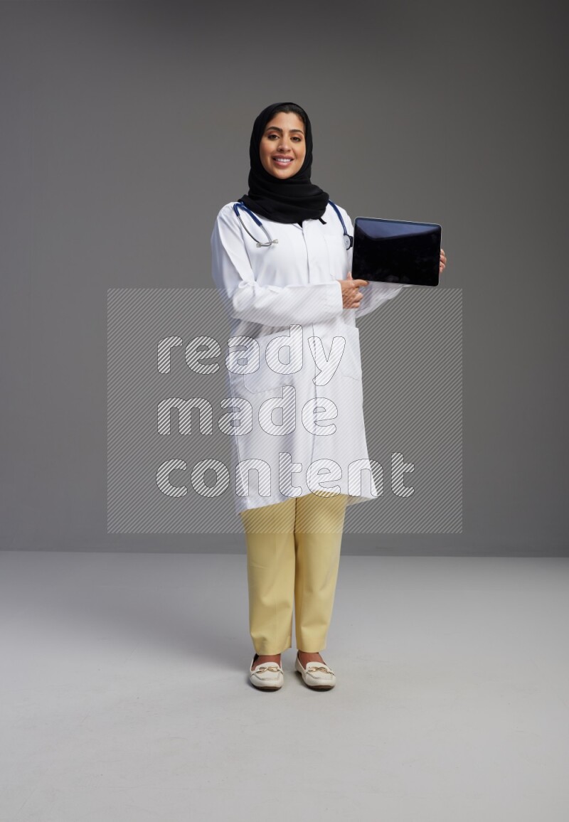 Saudi woman wearing lab coat with stethoscope standing showing tablet to camera with sign in the back on Gray background