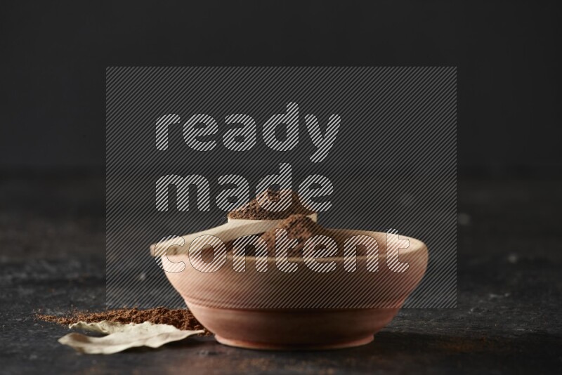 A wooden bowl and a wooden spoon full of cloves powder with laurel leaves on a textured black flooring