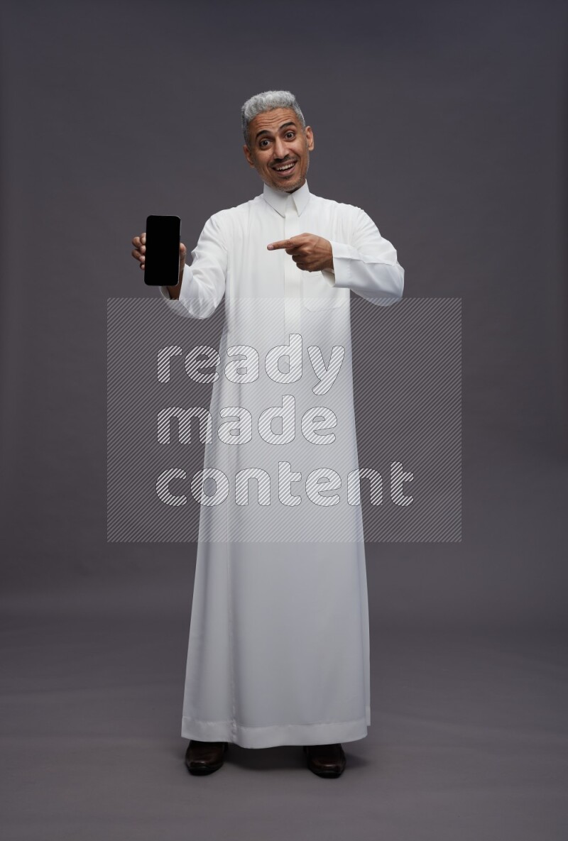 Saudi man wearing thob standing showing phone to camera on gray background
